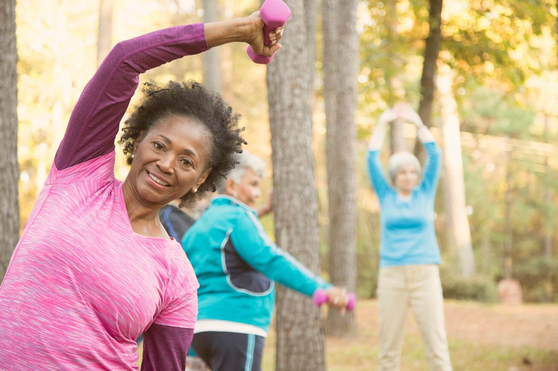 Senior African American woman smiling while exercising with weights in a park with other seniors in background, promoting active lifestyle for elderly homecare