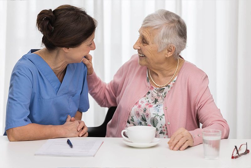 Caregiver sitting at a table with a elderly lady providing companion care and smiling